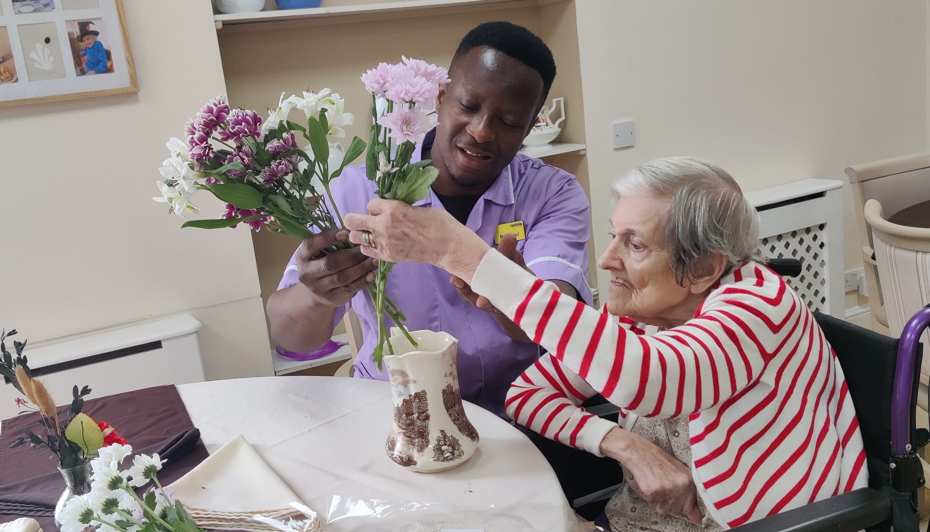 An older person in a wheelchair, wearing a red and white striped cardigan, arranges flowers in a vase with help from an Optalis staff member in a purple uniform. They are seated at a table with a white cloth, surrounded by flowers and items. Shelves and framed pictures are visible in the background.