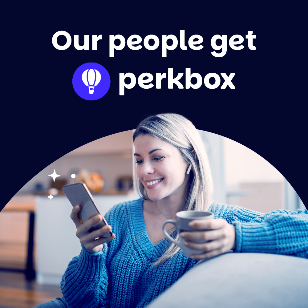 A person in a blue sweater sits on a couch, smiling while looking at their smartphone and holding a mug. Above them is the text "Our people get Perkbox" with a hot air balloon icon. The background is a softly lit indoor setting.
